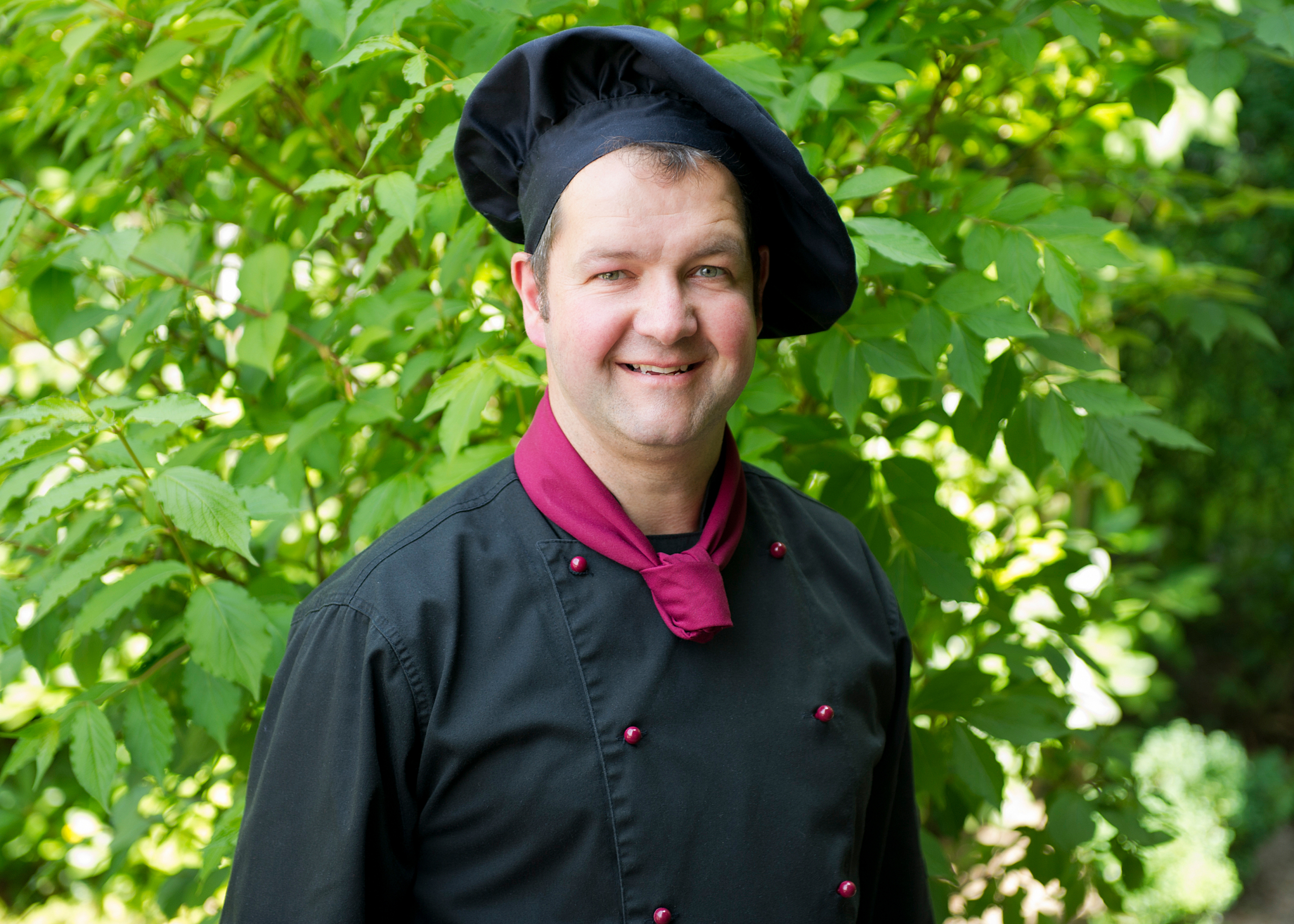 Smiling chef in a black hat and jacket with a burgundy neckerchief stands among sunlit green foliage.